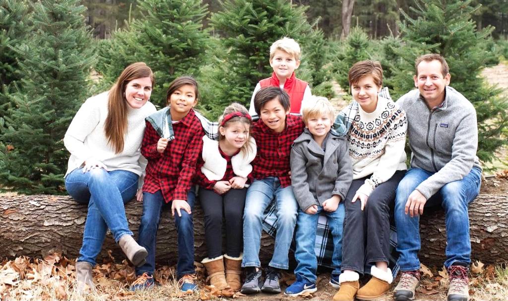 Christy Lillie with her husband and their six children, seated together on a log at a Christmas tree farm.